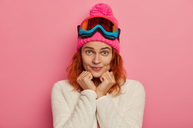 Attractve redhead woman wears warm hat, ski goggles and white jumper