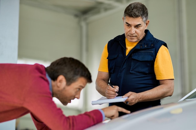 Free photo auto repair man writing notes while his customer is pointing at problematic spot on vehicle hood in a workshop