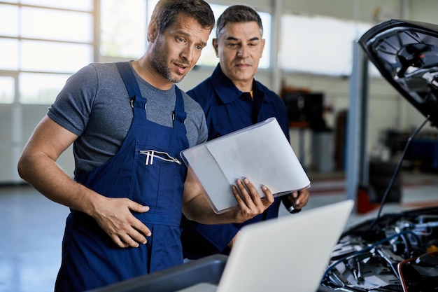 Auto repairman and his coworker using laptop while analyzing car's performance in a workshop