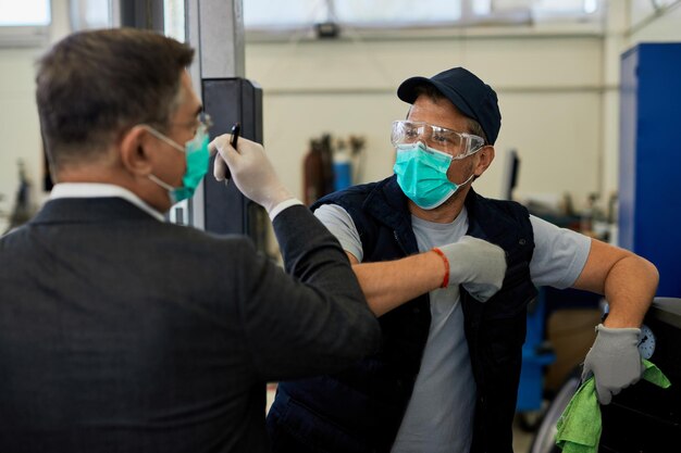 Auto repairman and his manager wearing protective face masks and greeting with elbows in a workshop