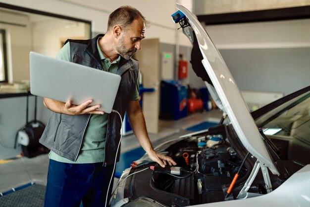 Auto repairman using laptop while examining car engine in a workshop