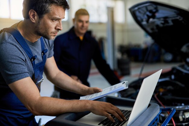 Auto repairman working on a computer while doing car diagnostic with his coworker in a workshop