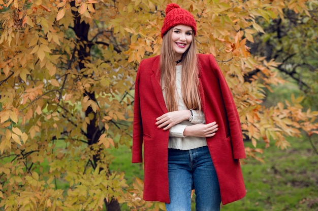 Free photo autumn fashion portrait of happy blonde woman in red stylish coat and knitted hat walking in park.