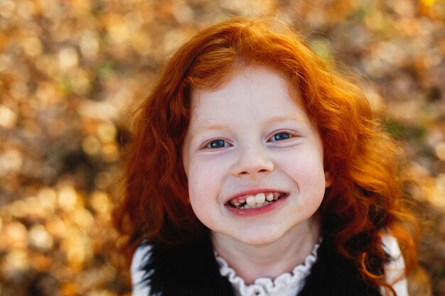 Autumn vibes, child portrait. Charming and red hair little girl looks happy standing on the fallen l