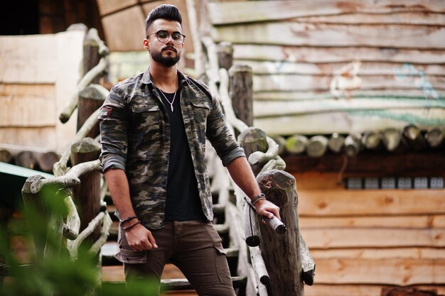 Awesome beautiful tall ararbian beard macho man in glasses and military jacket posed outdoor against wooden stairs