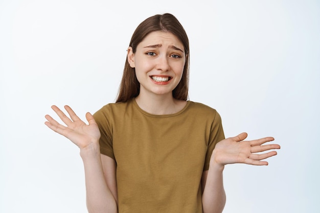 Awkward girl shrugging shoulders with apologizing face expression, looking clueless, say sorry, showing empty hands, has nothing, standing over white background.