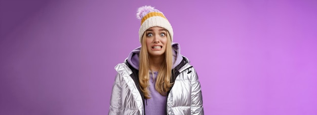 Free photo awkward worried cute timid blond girl in silver jacket hoodie winter hat clench teeth popping eyes camera ooops make mistake standing nervous someone notice purple background