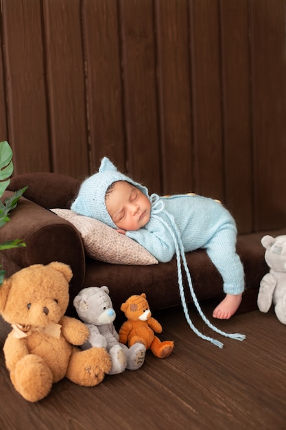 Baby boy little newborn resting on brown sofa in blue crocheted pijamas surrounded by cute toy bears