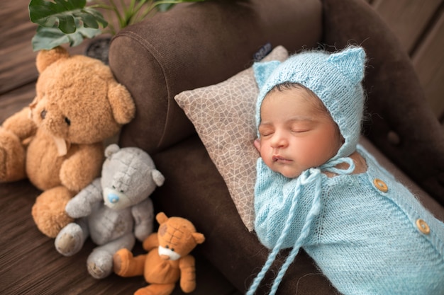 Baby boy newborn baby sleeping in blue crocheted pijamas on brown sofa surrounded by three toy bears