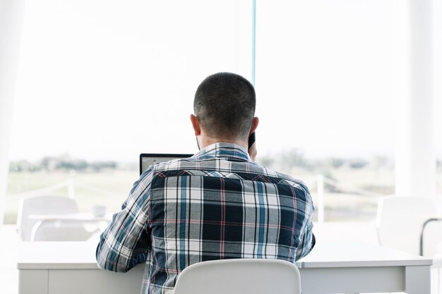 Back of a man working in front of the laptop and talking to the mobile