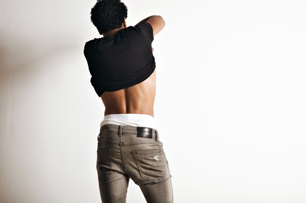 Back portrait of a muscular young model taking off his black unlabeled short sleeve t-shirt on white wall