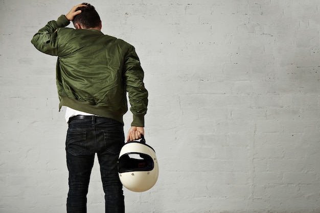 Back portrait of a young motorcyclist in jeans, military bomber jacket and holding his white helmet touching his hair isolated on white