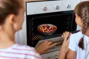 Free photo back side portrait of young woman putting croissant to baking dish of oven, her daughter standing near by and looking inside gas-stove, wants to taste delicious as soon as possible.