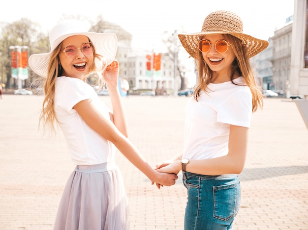 Free Photo back of two young beautiful blond smiling hipster girls in trendy summer white t-shirt clothes and hat.  . couple holding each other hands