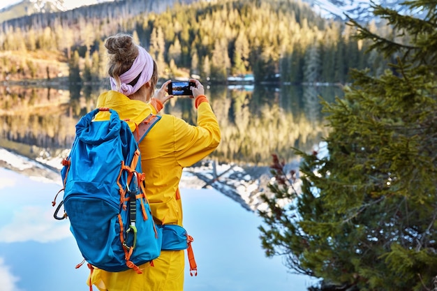 Back view of active female tourist photographs lakescape with mountains on her smart phone device