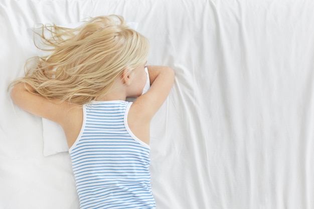 Free photo back view of adorable blonde girl wearing striped t-shirt, having healthy sleep, lying on stomach on white pillow, dreaming about something. restful carefree small kid sleeping in bed after school