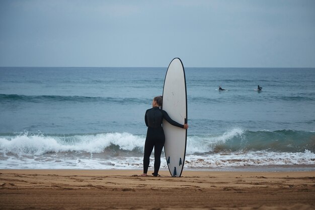 Back view on beautiful young surf girl hugging her longboard on ocean shore and watching waves before surfing