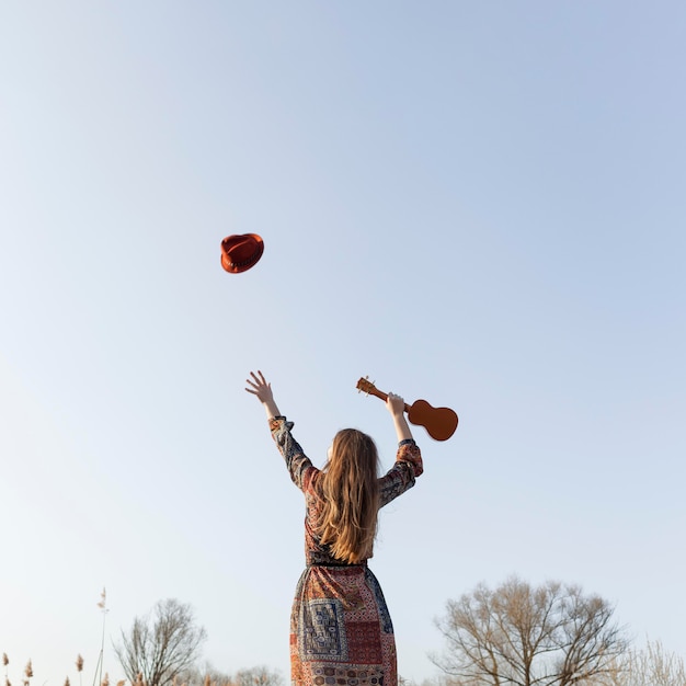 Back view of bohemian woman throwing hat in the air and holding ukulele