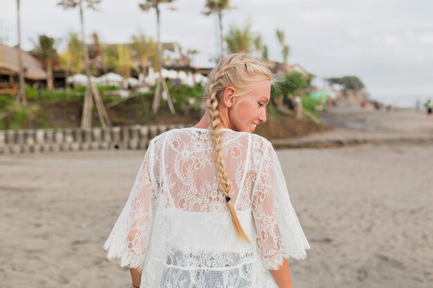 Back view of charming woman with blond in white blouse walking down the beach