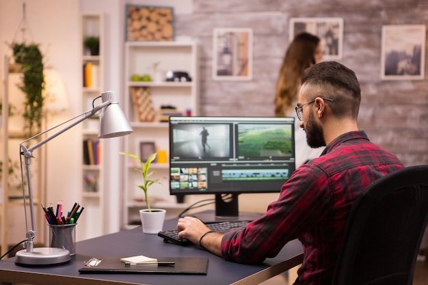 Back view of creative filmmaker working on a movie on laptop. Girlfriend in the background.