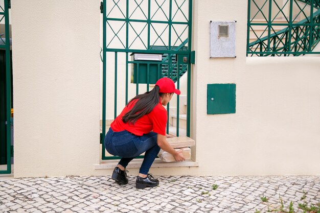 Back view of female courier putting parcels near gate. Long-haired brunette deliverywoman in red uniform squatting and delivering express order to customer at home. Delivery service and post concept