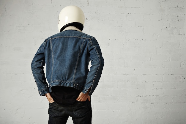 Back view on fit body of younf motocyclist wears helmet, black longsleeve henley shirt and club denim jacket with his hands in back pockets of his trousers