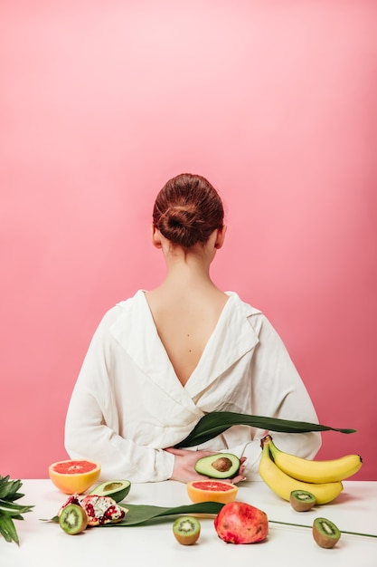 Free photo back view of ginger woman with exotic fruits. studio shot of girl in white shirt with bananas, grapefruits, garnet. kiwi and avocado on pink background.