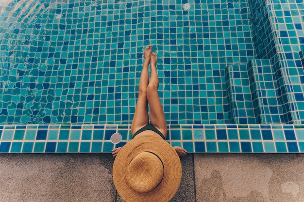 Back view of graceful woman in swimsuit and hat sitting near the pool