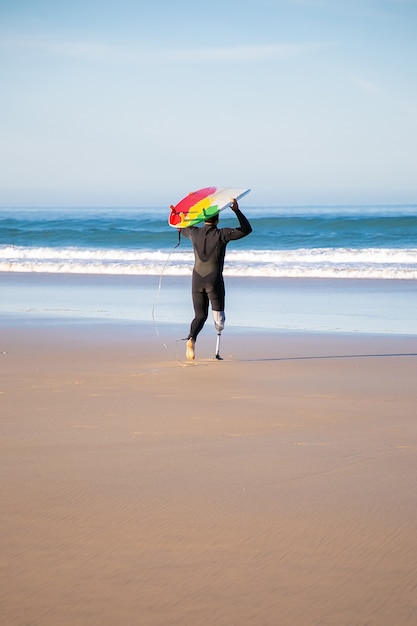 Free photo back view of handicapped surfer going to sea with board. active man with amputated leg holding surfboard and surfing at summer vacation