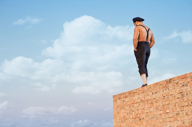 Free photo back view of muscular builder in work wear standing on brick wall on high. man holding hands in pockets and looking down. extreme building in hot summer day. blue sky on background.