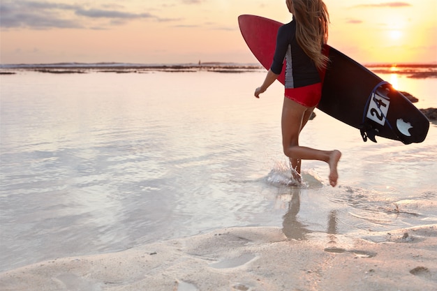 Back view of running girl surfer in swimsuit, carries board under arm, ready to conquer giant wave, runs into ocean