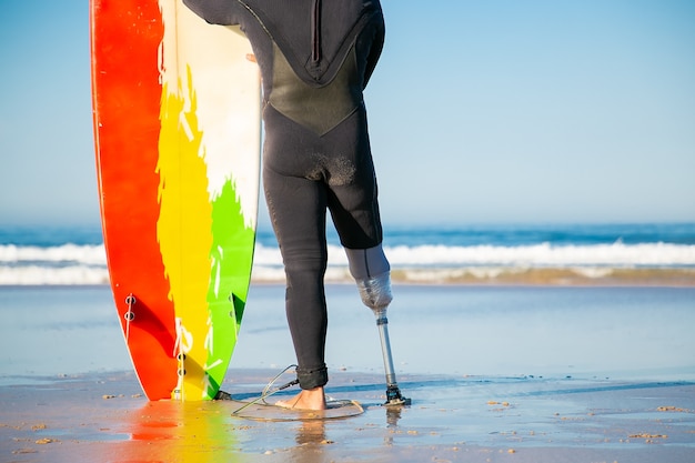 Free photo back view of unrecognizable amputee standing with surfboard on beach