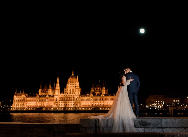 Back view of wedding couple in love with picturesque illuminated parliament in Budapest in the night