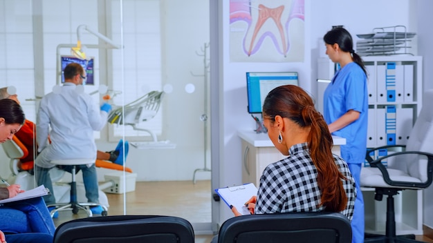 Back view of woman filling in dental document sitting on chiar in waiting room preparing for teeth exemination while doctor working in background. Concept of crowded professional orthodontist office.