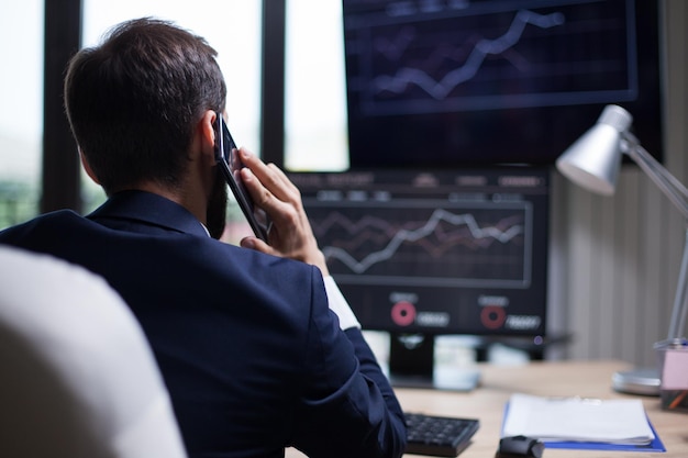 Back view of young business bearded man having a phone call with his CEO. Diagrams on computer monitor.