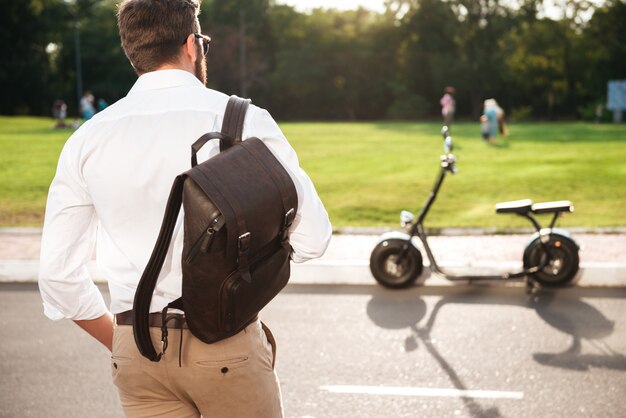 Back view of young man with backpack posing near the modern motorbike outdoors