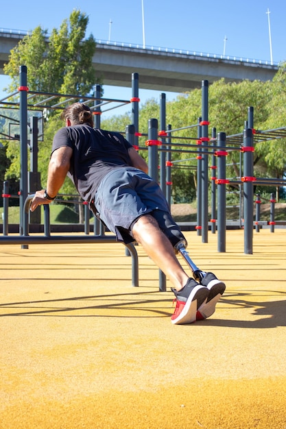 Back view of young man with prosthetic leg doing push-ups. Sporty man doing sports on stadium training hands leaning on bar over ground. Motivation for active life for people with disability concept