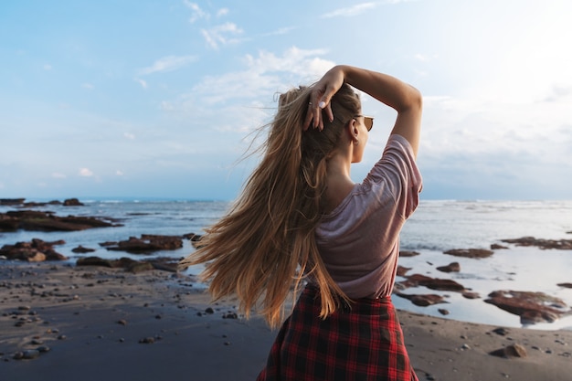 Backside view a traveller girl with long hair, enjoying blue ocean view standing black volcanic sand beach.