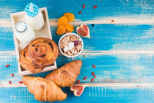 Baked breads with milk bottles in tray near dry apricots; fig fruit; and corn flakes over blue wooden background