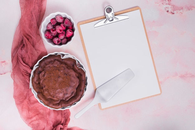 Baked cake; raspberry; spatula on clipboard with white paper over the pink textured background