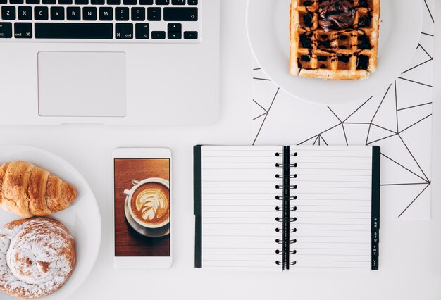 Baked food; chocolate waffle; mobile phone with coffee screen; laptop and spiral notepad on white desk