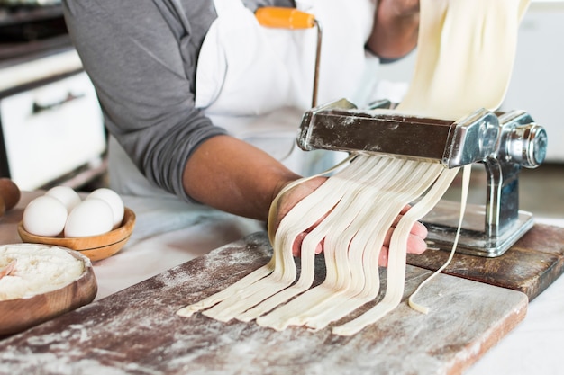 Baker cutting raw dough into tagliatelle on pasta machine over the wooden board