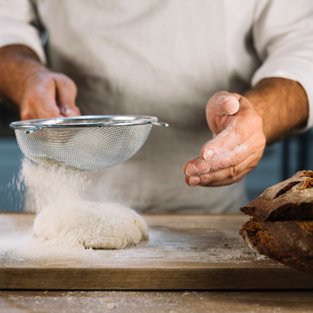 Free photo baker sifting the wheat flour through steel sieve over knead dough