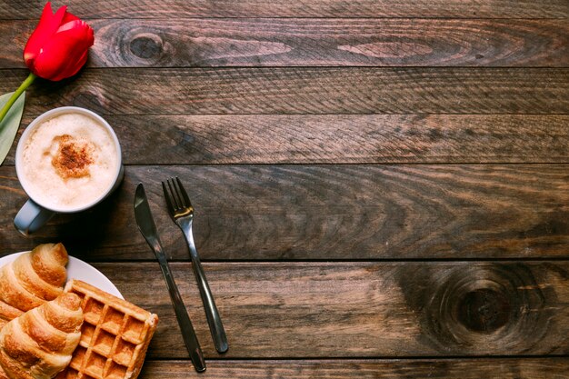 Bakery on plate near cup of drink, flower and cutlery