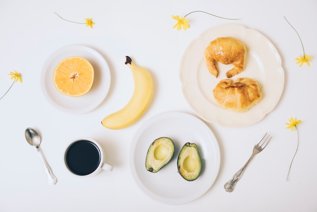 Banana; croissants; halved avocado; coffee cup on white backdrop with spoon and fork on white backdrop