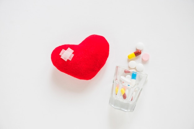 Bandage on red heart stuffed with many pills spilling from glass on white background