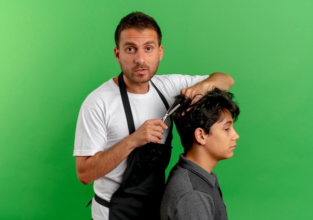 Free photo barber man in apron cutting hair with scissors of satisfied client standing over green wall