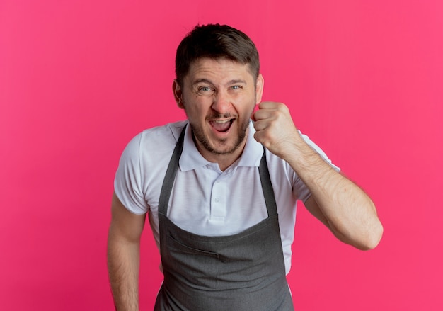 barber man in apron  going wild clenching fist shouting with aggressive expression standing over pink wall