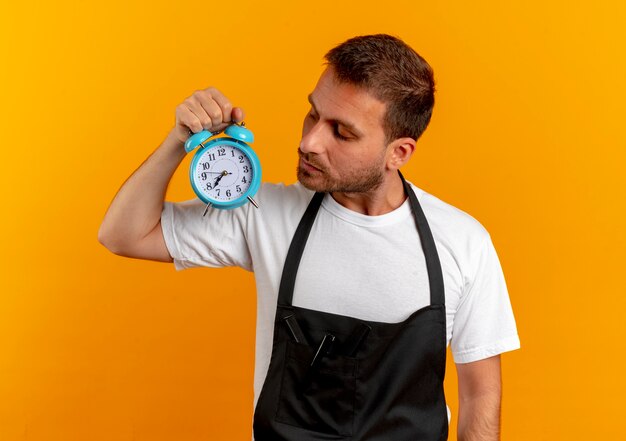 Barber man in apron holding alarm clock looking at it with serious face standing over orange wall