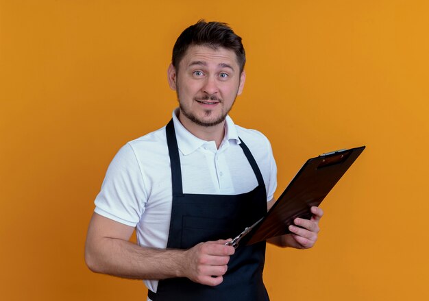 Barber man in apron holding clipboard and scissors looking at camera with smile on face standing over orange background